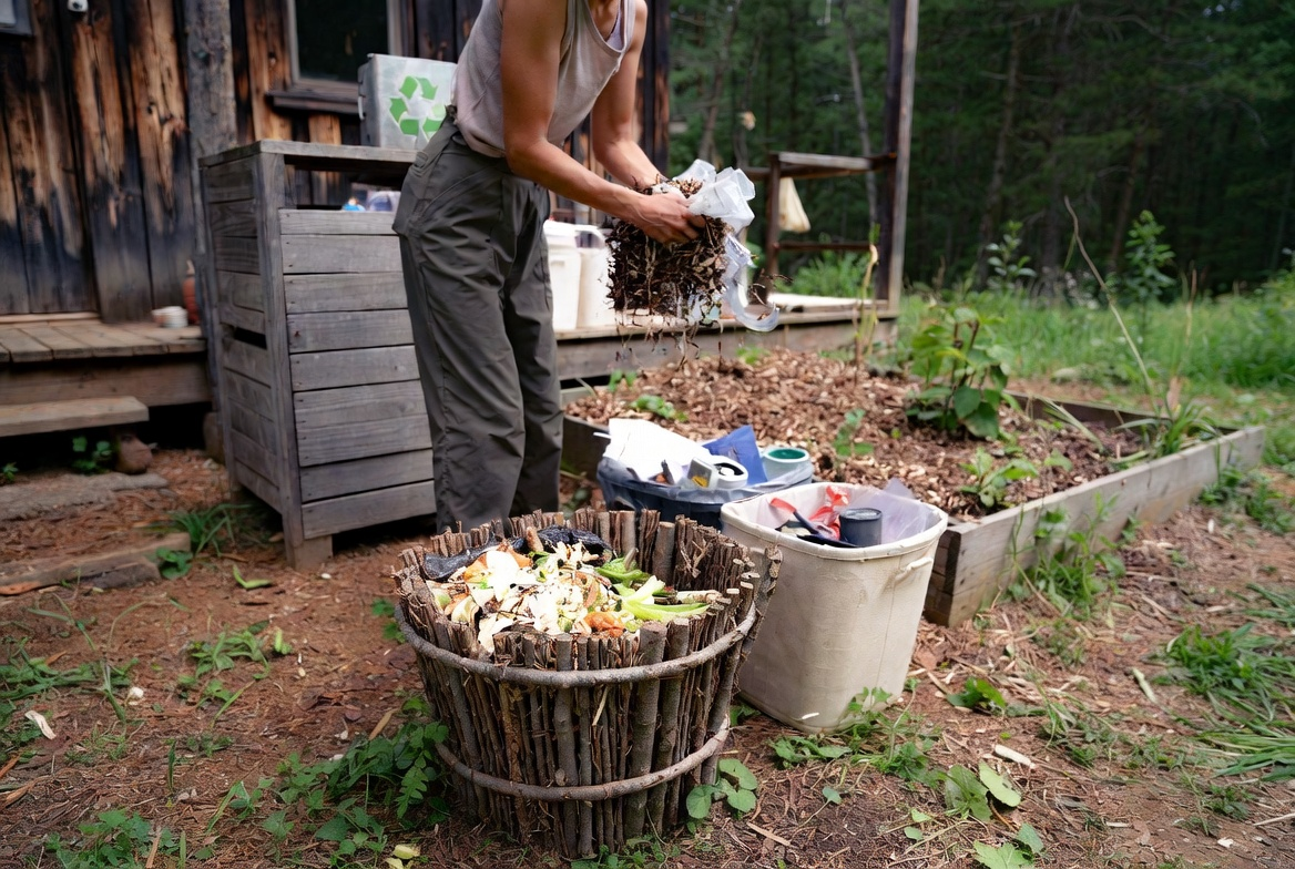 Homesteader adding kitchen scraps to a large wooden compost barrel outdoors at an off-grid homestead. Blue recycling bins and beige trash bin nearby, with a rustic wooden shed and forest in the background.